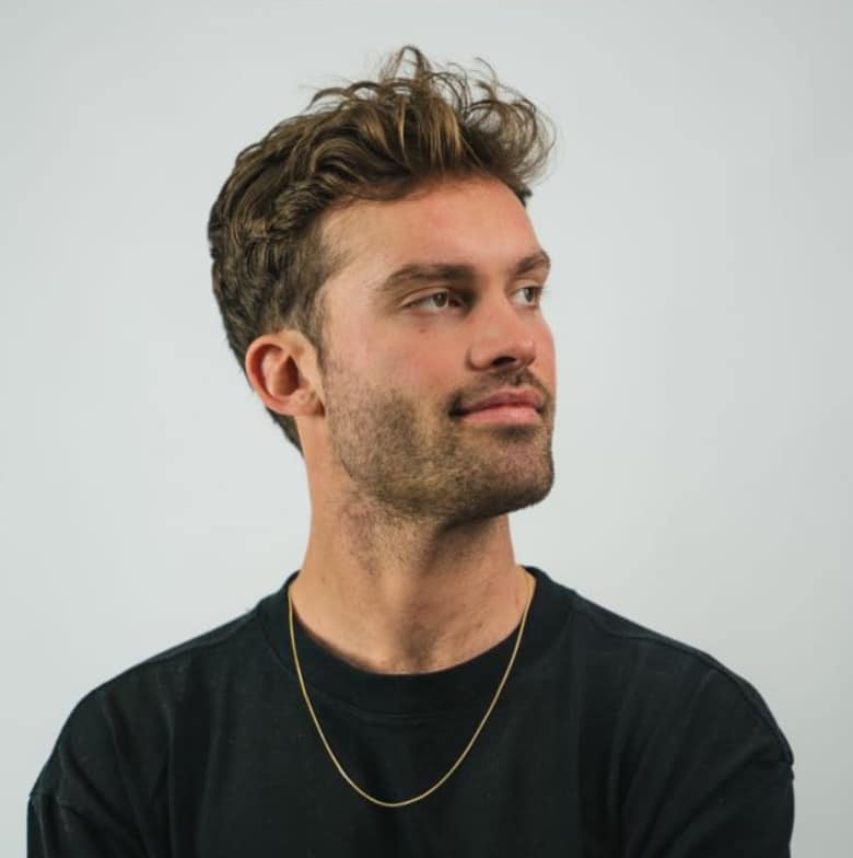 Een man genaamd Davy, met golvend bruin haar, een korte baard en een zwart shirt, kijkt opzij tegen een saaie achtergrond.